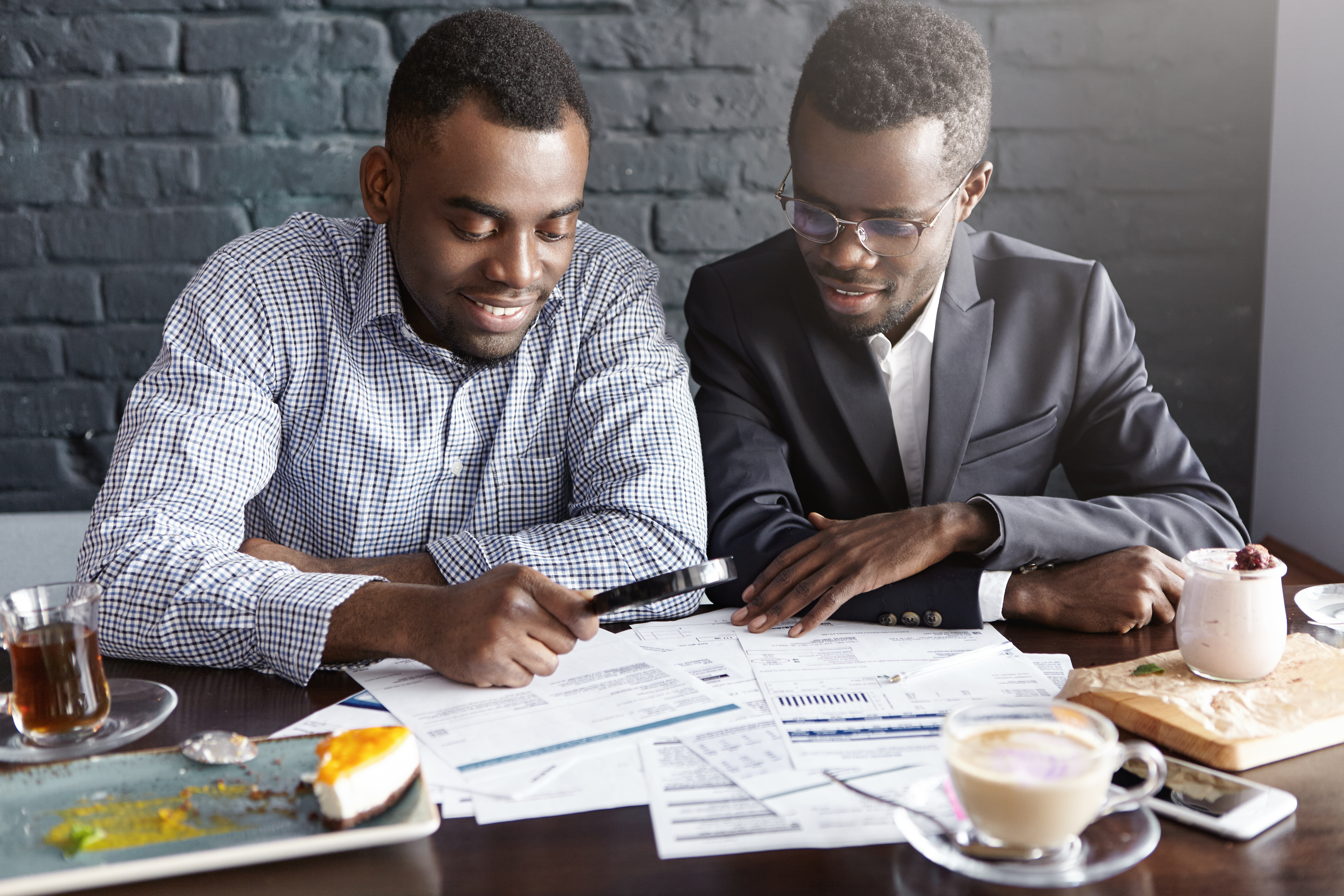 Two handsome successful African-American businessmen using magnifying glass while studying documents and reviewing finances, sitting at cafe table with papers, having happy cheerful expressions